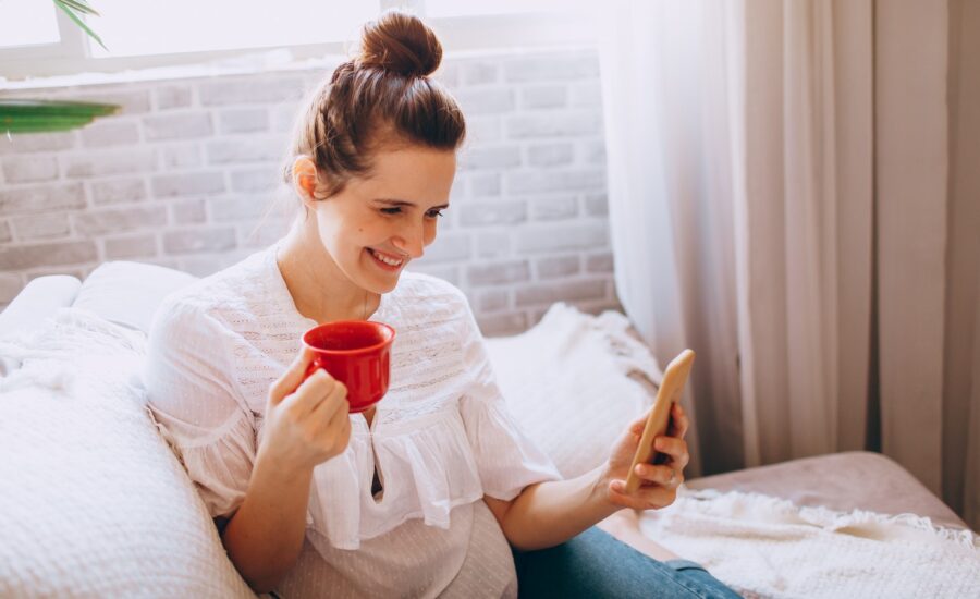 A woman drinks tea on a sofa, smiling at her phone