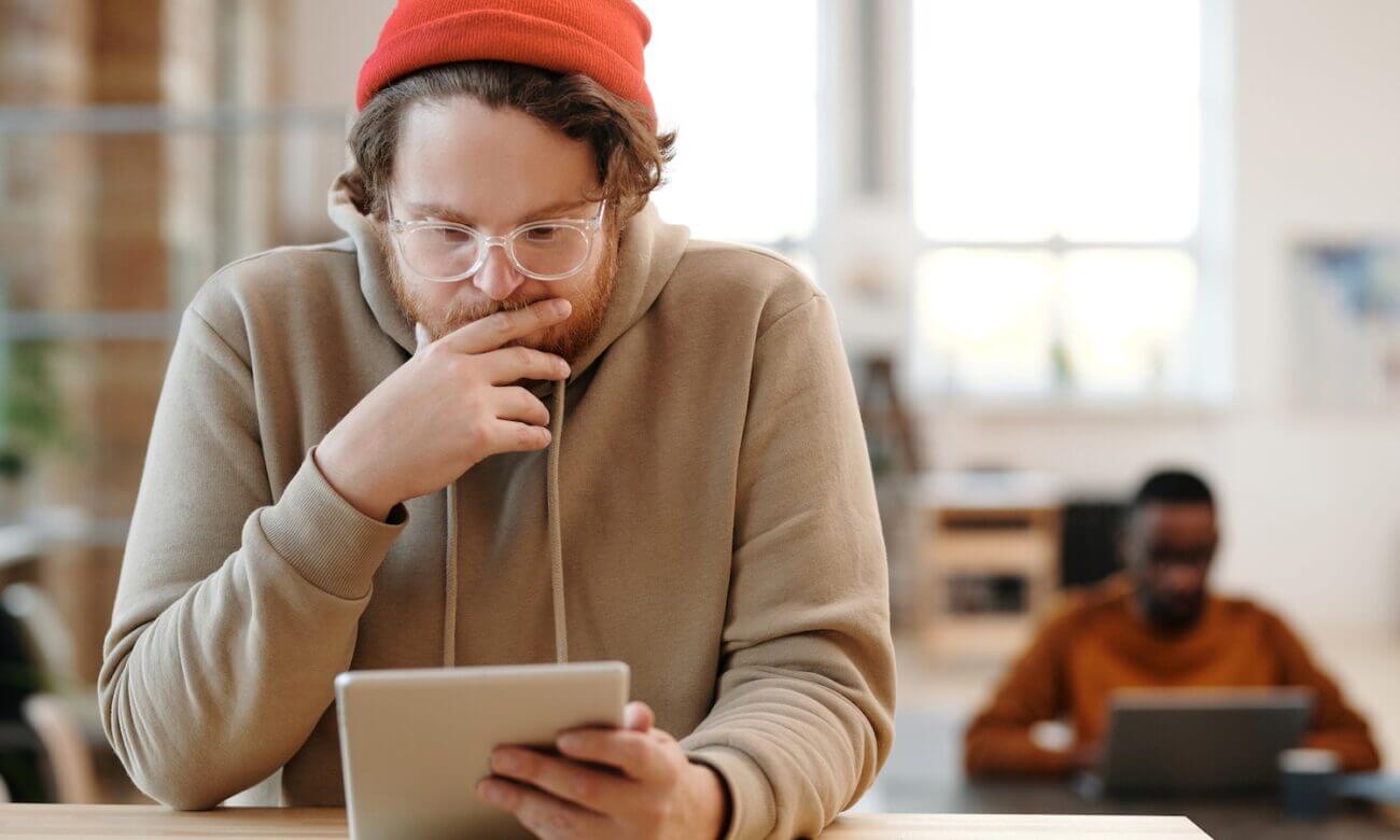 A man in a hat looks at something on his tablet with a thoughtful expression while sitting at a table.