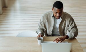 A man sits at a wooden desk and holds a cup of coffee while browsing on his laptop.