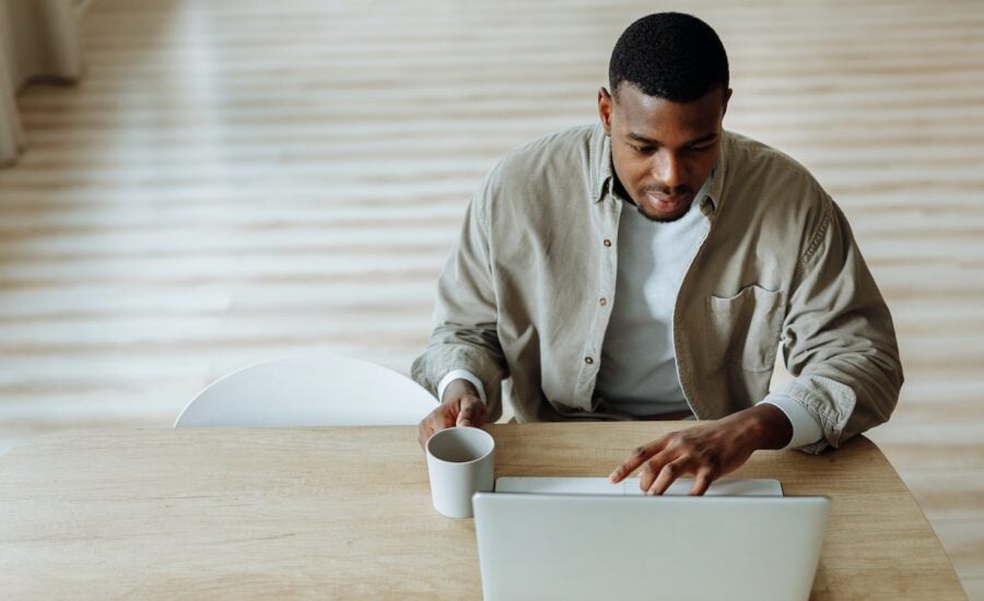 A man sits at a wooden desk and holds a cup of coffee while browsing on his laptop.