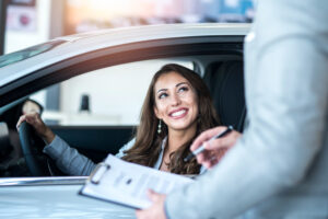 A woman sits behind the wheel of a car, smiling at a salesperson