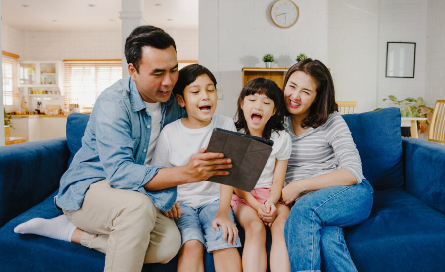 Parents and two young kids sit on a sofa and watch a tablet