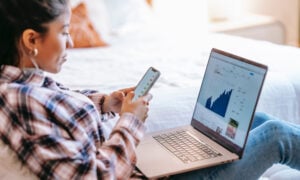 A woman sits in her bedroom, looking at her investments both on her phone and her laptop