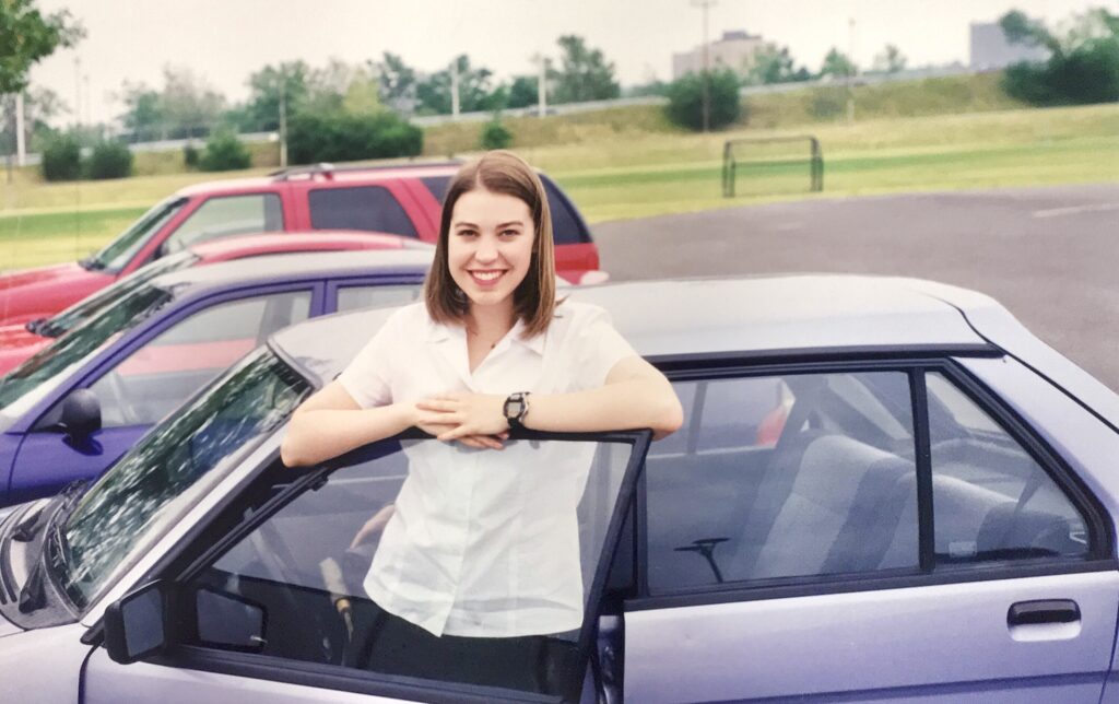 A smiling young woman beside a small, purple Subaru Justy car
