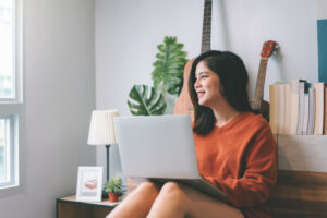 A smiling young woman sits with her laptop and looks out the window