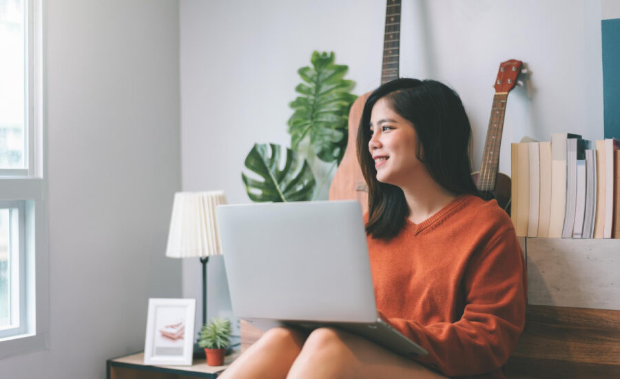 A smiling young woman sits with her laptop and looks out the window