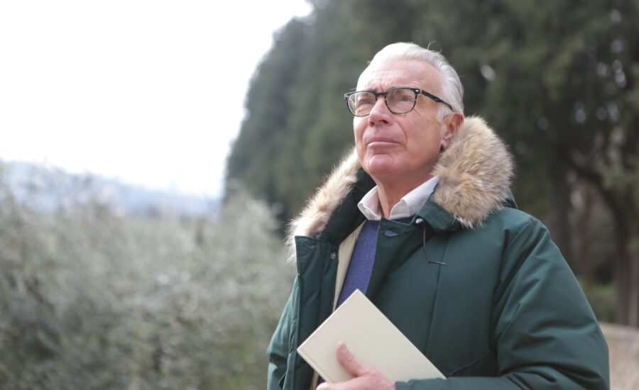An older man in a winter coat stands in a wooded area and looks worried