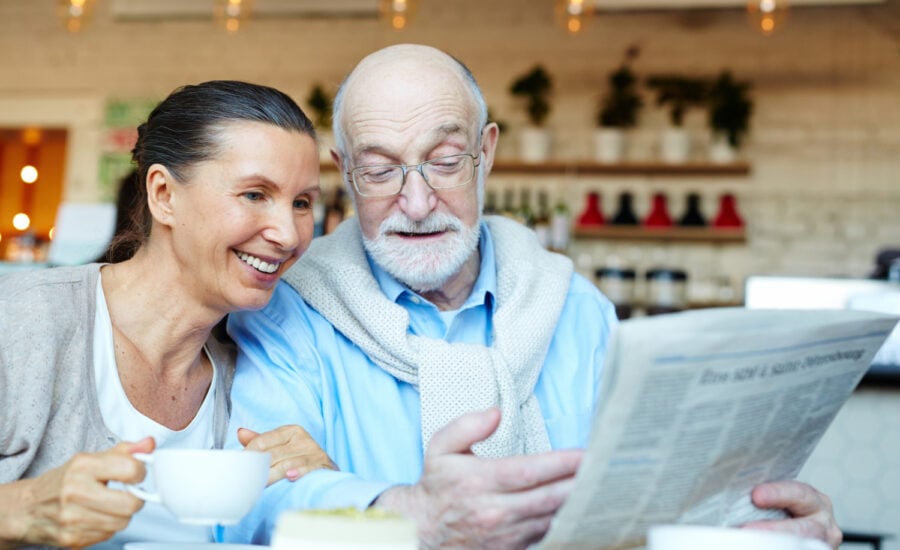 A smiling senior-age couple in a cafe discuss a newspaper article