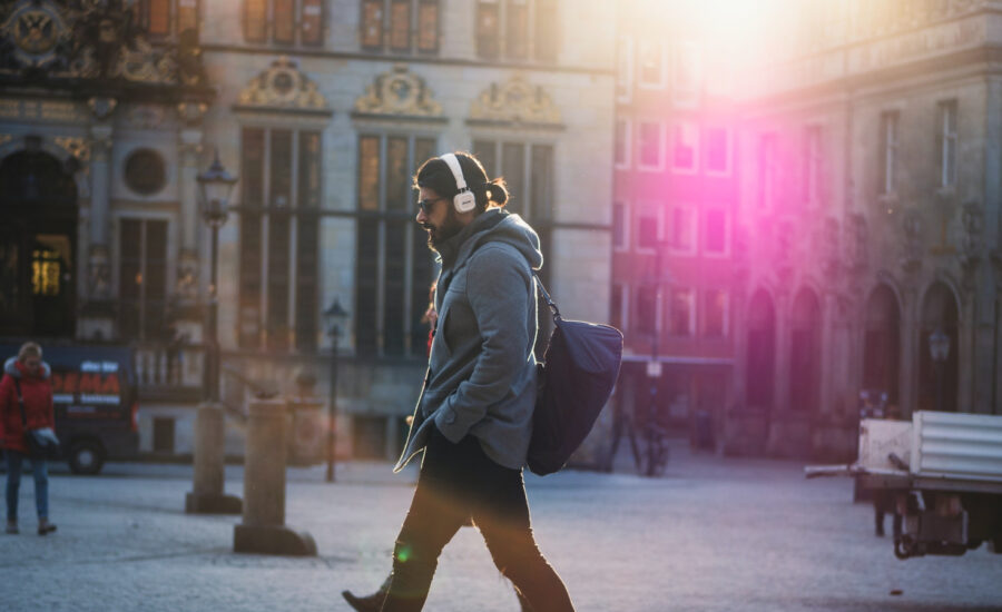 A man walks along a European street wearing headphones and carrying a bag