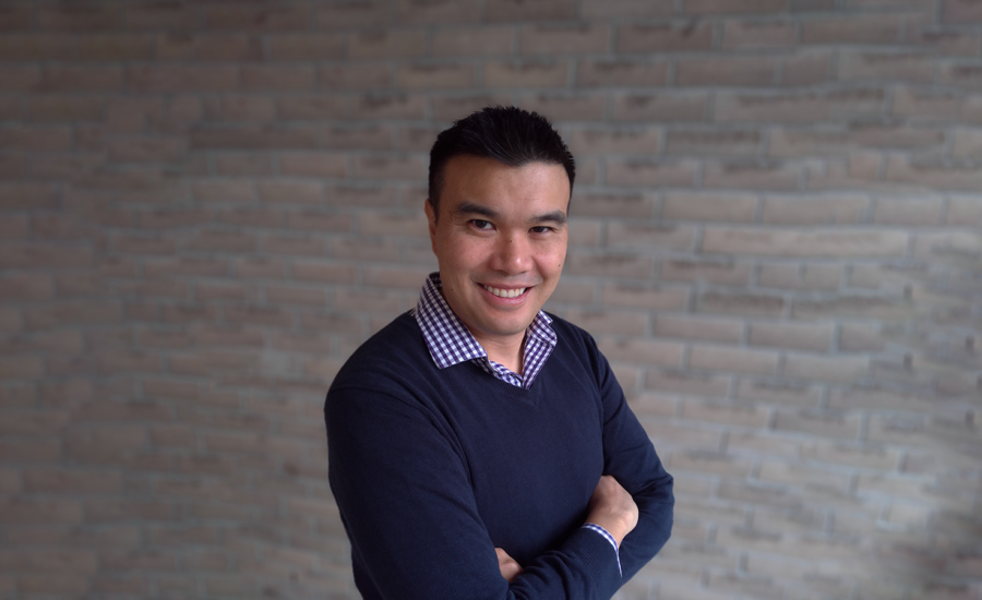 Certified Financial Planner Nicholas Hui smiles at the camera while crossing his arms with a grey brick wall in the background.