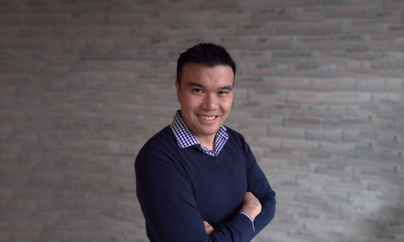 Certified Financial Planner Nicholas Hui smiles at the camera while crossing his arms with a grey brick wall in the background.