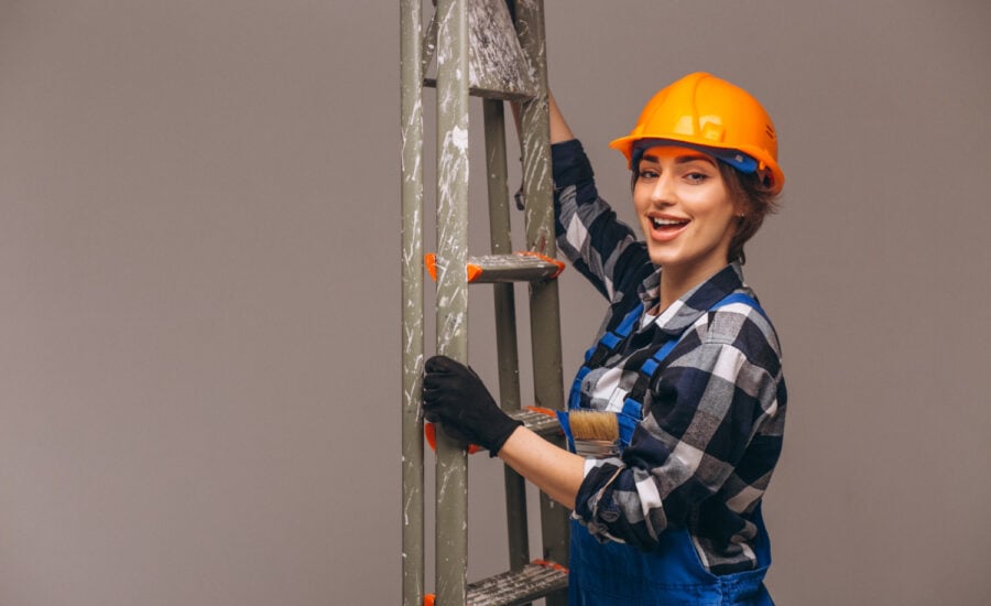 A smiling woman in work clothes holds a ladder