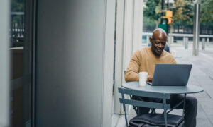 A man sits outside of a coffee shop, working on his laptop