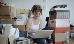 A woman sits on the floor, surrounded by boxes of her belongings, to illustrate how to make money selling stuff online.