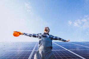 A man in work clothes stands in front of a solar panel array and opens his arms to the sun.