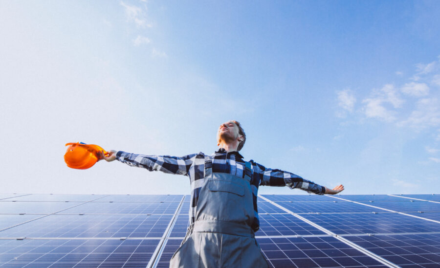 A man in work clothes stands in front of a solar panel array and opens his arms to the sun.