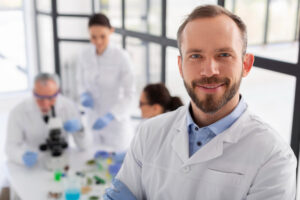 A smiling man in a lab coat, in front of three other scientists at a table