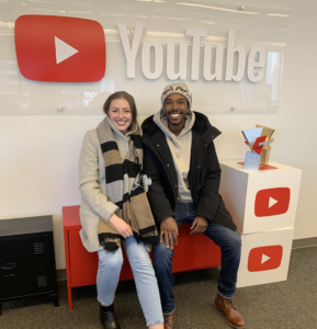Steph and Den sit in front of a YouTube sign on a red couch at YouTube's head office in Canada.
