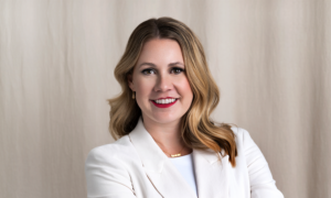 The Pink Tax author Janine Rogan sits on a white cabinet while smiling slightly at the camera against a white background.