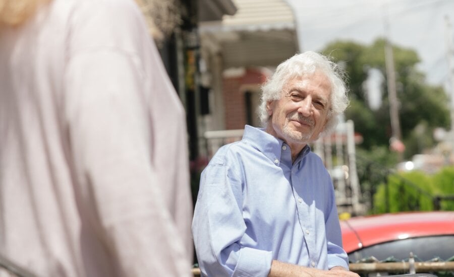 A smiling elderly man stands against a handrail in his driveway