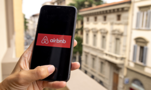 On a balcony on a European street, a hand holds a phone with Airbnb's logo