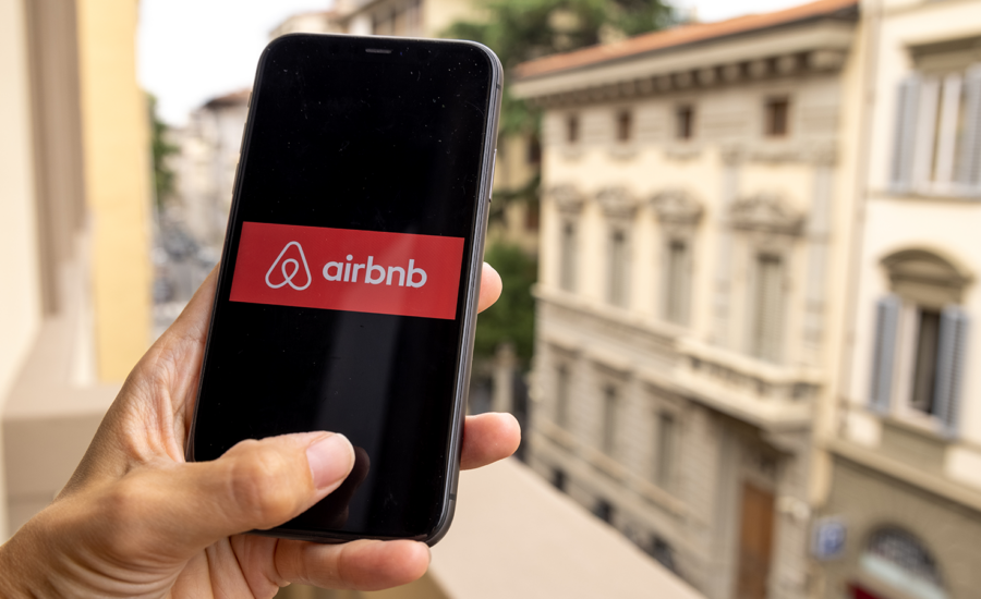 On a balcony on a European street, a hand holds a phone with Airbnb's logo