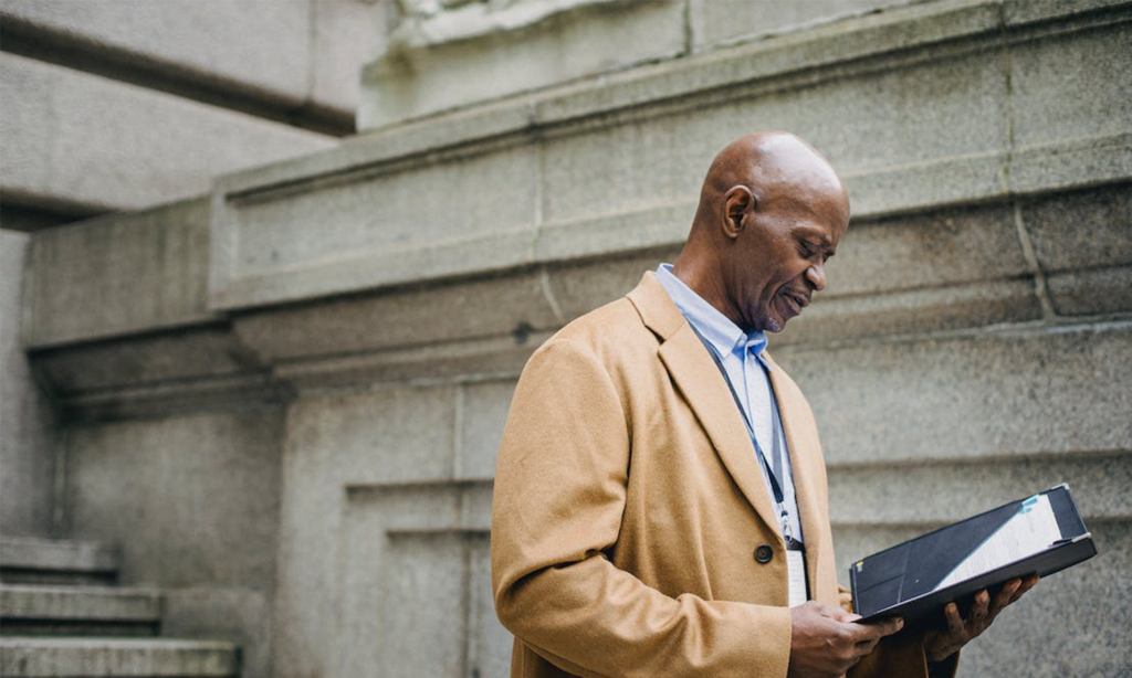 An employee in a beige suit stands outside looking at his pension plan summary in a folder.