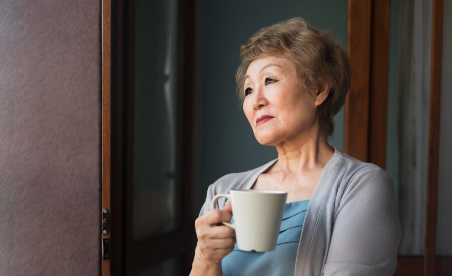 A middle-aged woman, holding a mug, gazes out the window