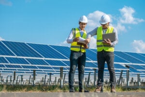 Two men in hard hats and safety vests stand in front of a solar panel array