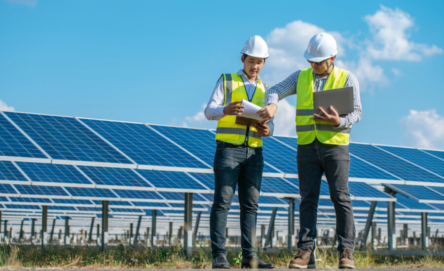Two men in hard hats and safety vests stand in front of a solar panel array