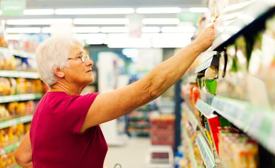 A senior-aged woman reaches for a product on a supermarket shelf