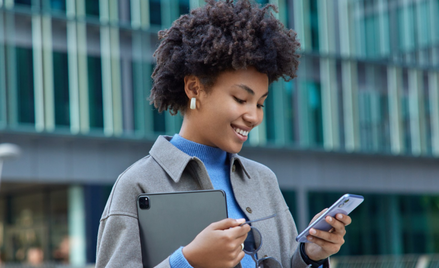 A smiling young woman looks at her phone on a downtown street.