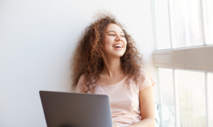 A smiling young woman holds her laptop beside a classroom window