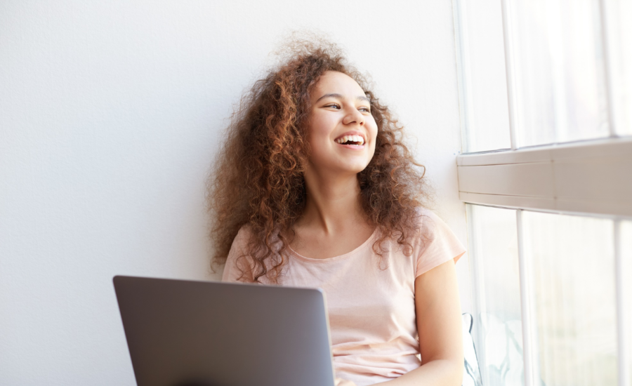 A smiling young woman holds her laptop beside a classroom window