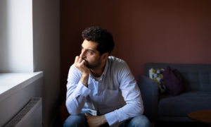 A worried-looking man looks out the window of his living room