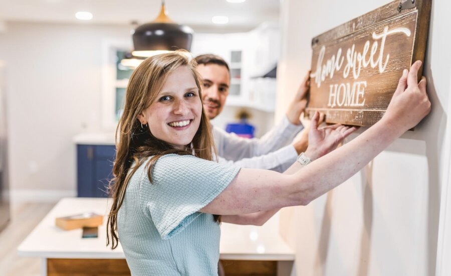 A young couple hangs a "home, sweet home" sign in their new condo