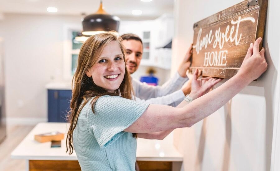 A young couple hangs a "home, sweet home" sign in their new condo