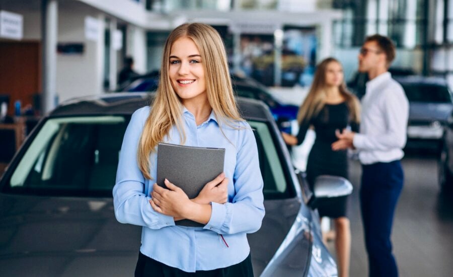 A young car saleswoman standing in front of a car and two customers