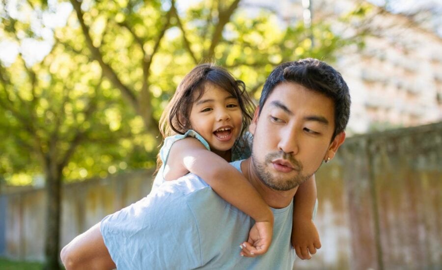 A young dad gives his toddler daughter a piggyback ride in the backyard