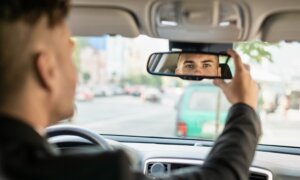 A car's rearview mirror reflects the eyes of a young man driving a car.