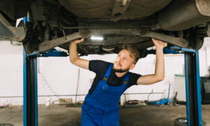 A man in coveralls stands below a hoisted car