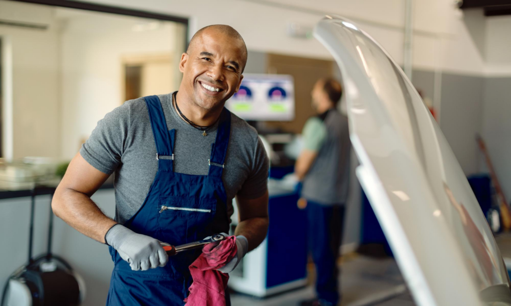 A smiling mechanic in front of a car with its hood up