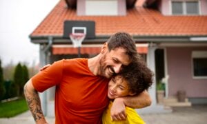 A dad and teenage son hug while playing basketball on their driveway
