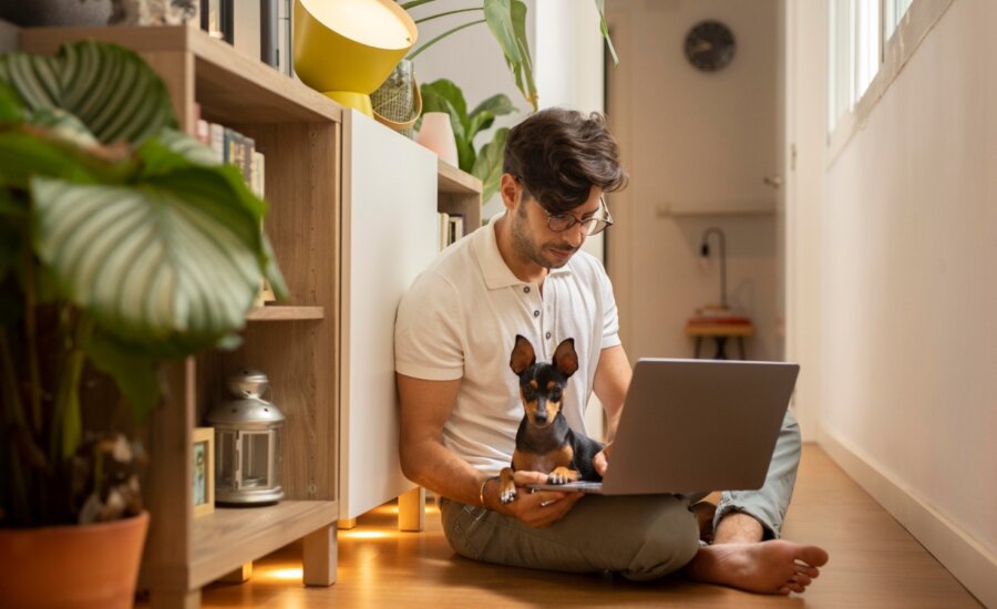 A man works on his laptop with a small dog on his lap