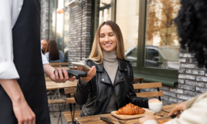 A woman taps her phone on a payment terminal on a cafe patio.