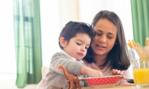 A toddler eats cereal while his mom watches