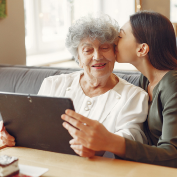 A young woman kisses her grandmother's cheek as they look at a tablet together