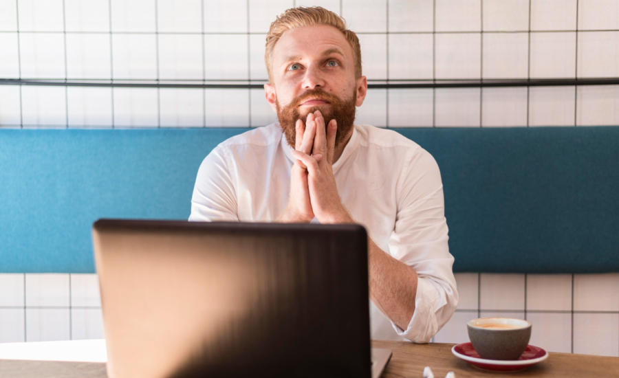 A man in a cafe with a laptop, looking up while thinking