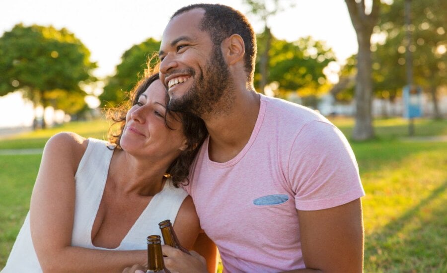 Happy couple enjoying outdoor date at sunset. Man and woman sitting on grass, talking, laughing, hugging and drinking. Romance concept