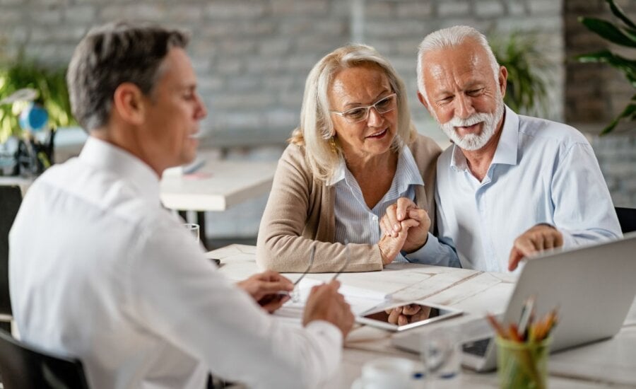 Happy senior couple holding hands and using laptop while having a meeting with financial advisor in the office. Senior man is pointing at something on laptop.